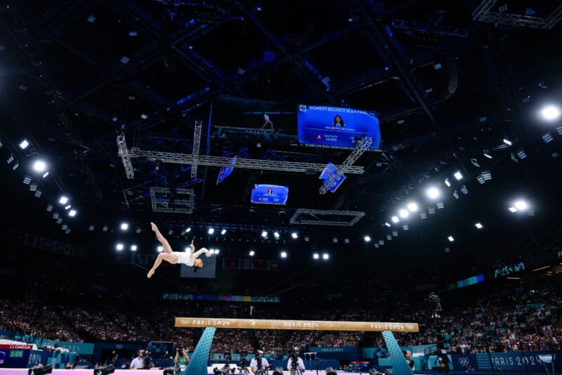 A gymnast mid-air performing a flip above the balance beam in a large, well-lit arena during an international competition. The audience in the stands watches closely, while screens and lights hang from the ceiling. Signs read "PARIS 2024.