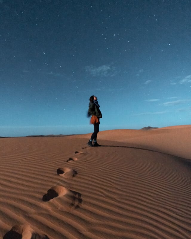 A person stands on a sand dune at night, looking up at a starry sky. The desert landscape is illuminated by soft moonlight, with footprints visible in the sand leading to where they stand.