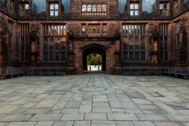 Gothic-style architectural structure with detailed stone carvings and arched doorways. The paved courtyard leads to an open archway revealing a glimpse of greenery beyond.