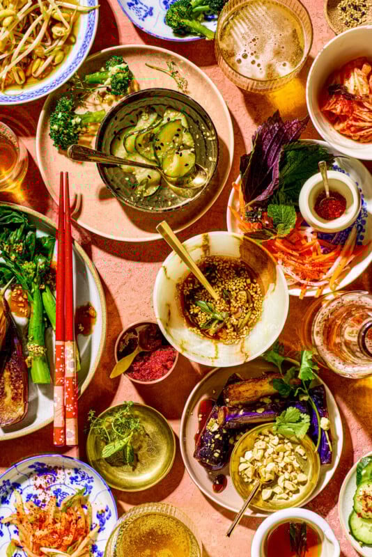 A vibrant display of various Asian dishes on a pink table. Plates are filled with pickles, kimchi, vegetables, spices, and garnishes. Red chopsticks and glasses add color and texture to the rich culinary spread.