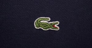 An embroidered green crocodile emblem with an open red mouth is sewn onto a textured dark blue fabric background.