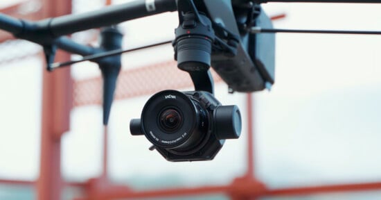 A close-up of a black camera mounted on a drone, with a blurred bridge structure in the background. the camera lens has a visible brand name.