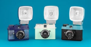 Three vintage-style cameras with flash attachments are displayed against a blue background. The cameras are colored dark blue, light mint, and black, each featuring a retro design with textured exteriors and prominent lenses.