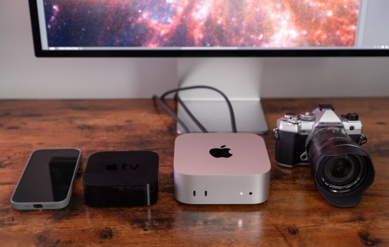 A smartphone, an Apple TV box, a Mac Mini, and a camera sit on a wooden desk. A large monitor displays a galaxy image in the background. Cables connect the Mac Mini to the monitor.