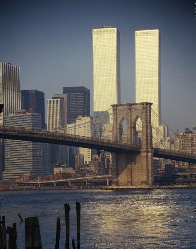 A view of the Brooklyn Bridge spanning the East River, with the Twin Towers of the World Trade Center rising prominently in the background among other skyscrapers in New York City. The scene is bathed in warm sunlight, reflecting off the water below.