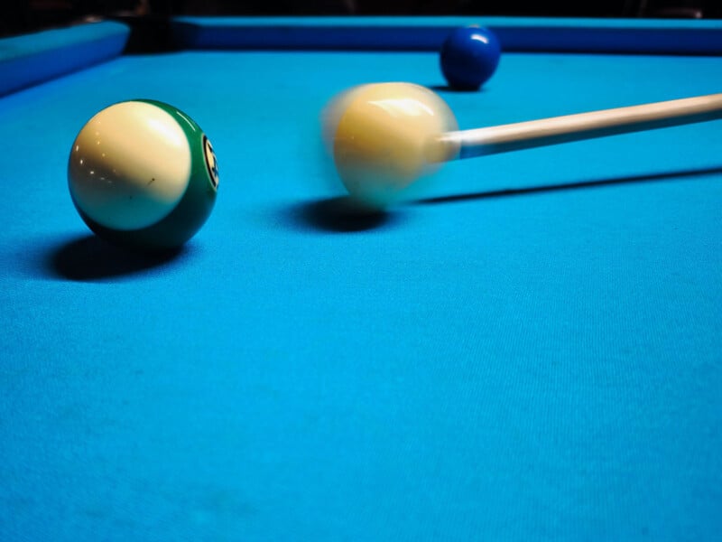A pool cue strikes a white cue ball on a blue pool table, sending it towards a green ball with the number six. A blurry motion effect is visible, capturing the moment of impact. A blue ball is visible in the background.